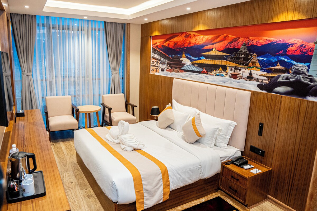 A hotel room with a large bed, two armchairs, a small table, a nightstand with a phone, and a painting of a mountain scene.
