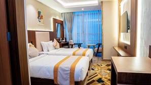 A hotel room with two beds, a desk, a chair, a TV, and a large window with curtains. - Grand Ambassador Business Hotel (Kathmandu)