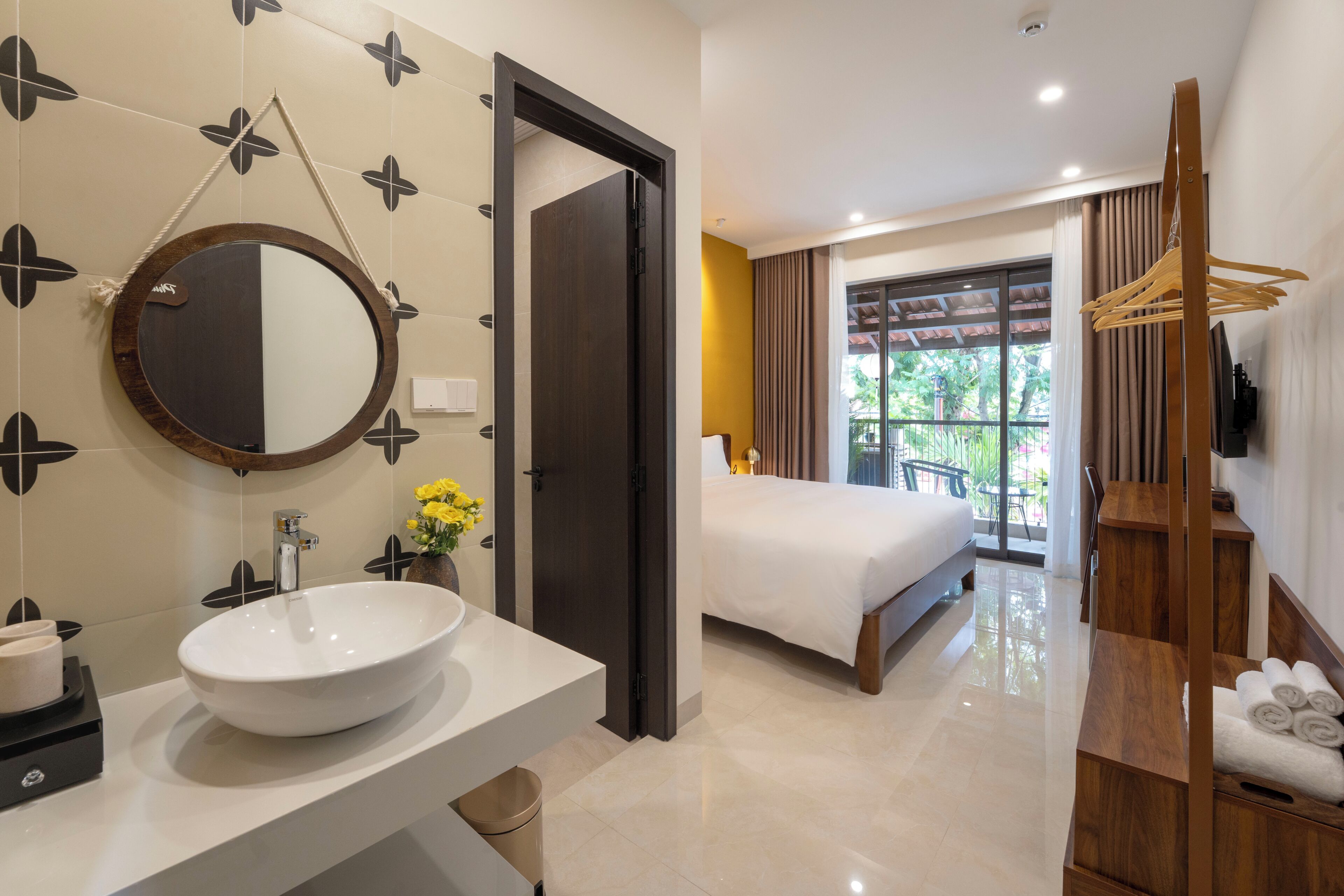 A modern hotel room with a large round mirror, a white sink, a bed, a wooden wardrobe, and a balcony with a view.