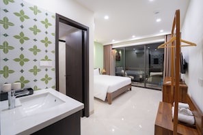 Habitación de hotel con baño, una cama, un perchero de madera y un ventanal corredero que da a un balcón.