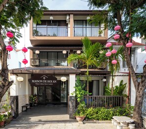En tvåvåningsbyggnad med en skylt som visar texten ”MAISON DE HOI AN” och en balkong med krukväxter.