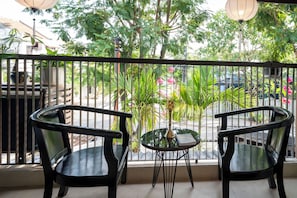 Balcón con dos sillas y una mesita, rodeado de barandilla metálica, con vistas a un jardín con diversas plantas y una calle.