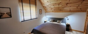 Une chambre avec un plafond en pente, une tête de lit en bois, un lit recouvert d’une couverture marron, deux lampes de chevet et un tableau encadré au mur.