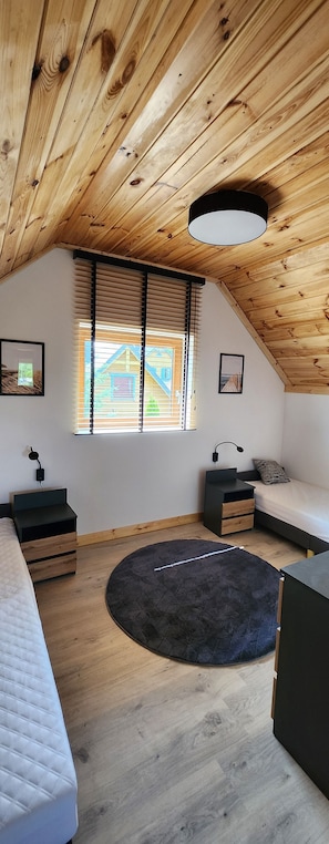 Un dormitorio con techo inclinado, vigas de madera, una cama, un escritorio, una silla, una alfombra redonda y una ventana con persianas.