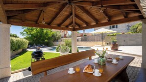 An outdoor patio with a wooden table set for breakfast, a grill, and a view of a swimming pool and garden. - Holiday house with pool for 10 persons, 2 separate accommodation units,in Porec (Dračevac)