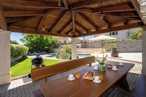 Eine Terrasse im Freien mit einem Holztisch, gedeckt für ein Frühstück, einem Grill sowie Blick auf ein Schwimmbad und einen Garten.