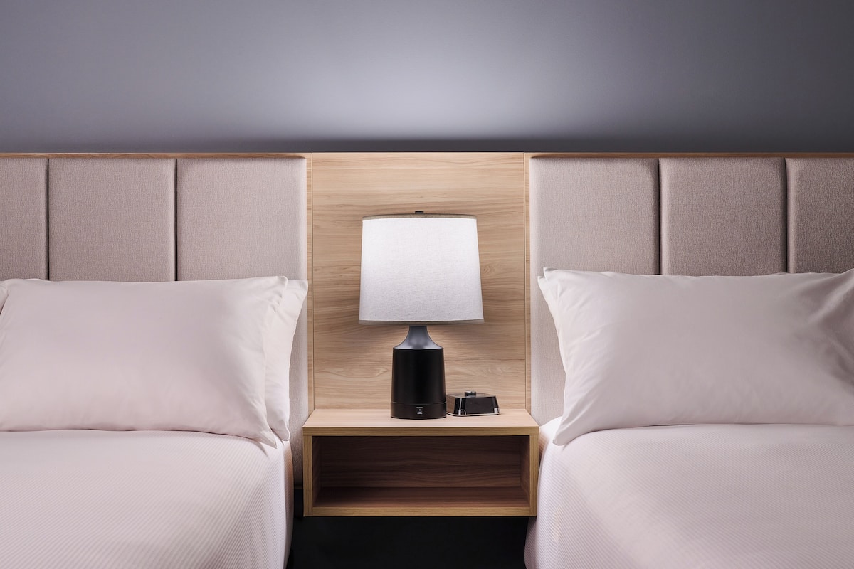 Hotellihuone, jossa on kaksi sänkyä, yöpöytä lampun kanssa ja puinen pääty.