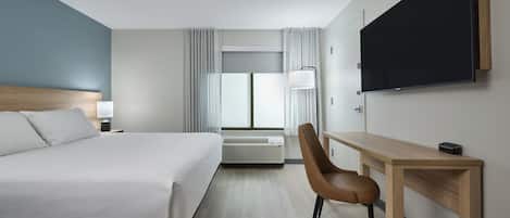 A hotel room with a bed, desk, chair, TV, and air conditioning unit.