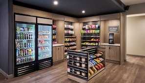 A self-checkout area with a 24/7 market, refrigerated beverage dispensers, and shelves stocked with snacks and beverages.