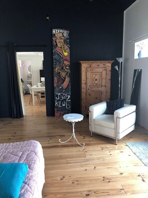 Ein Zimmer mit einem Bett, einem weißen Stuhl, einem kleinen weißen Tisch, einem Holzschrank und einer Wand mit einem farbenfrohen Wandgemälde.