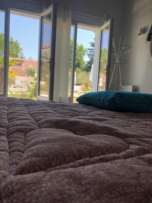 Ein Schlafzimmer mit einem geblümten Bettbezug, einem türkisblauen Kissen und Blick auf einen Garten durch eine Schiebetür.
