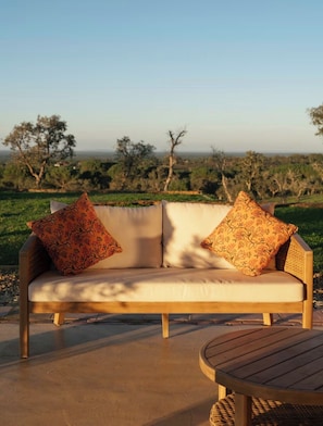 Ein Sofa mit floralen Kissen, ein runder Holztisch und Blick auf eine grüne Landschaft mit Bäumen.