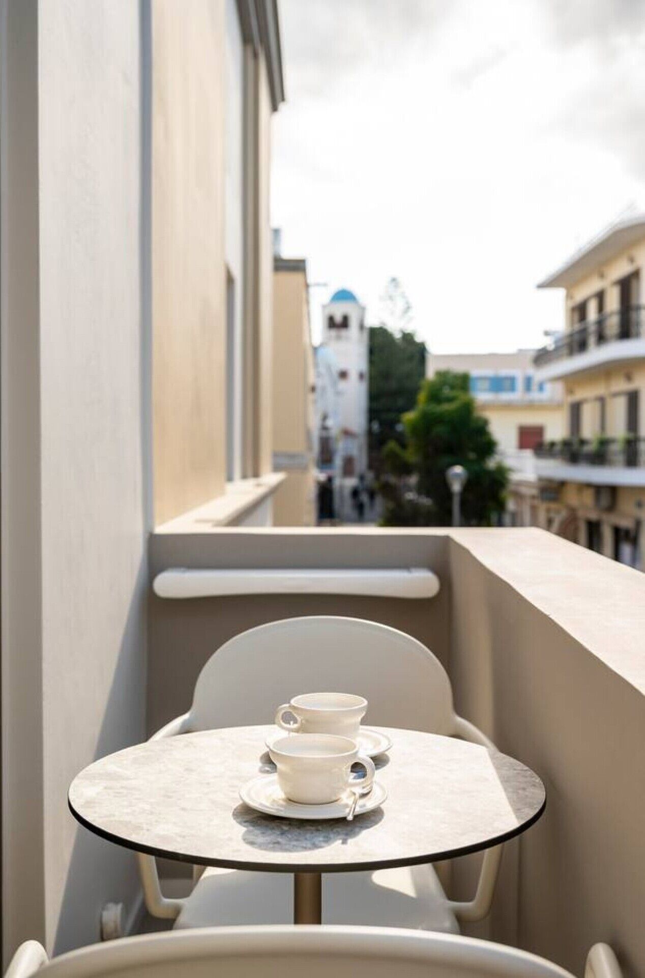 Un balcó amb una taula petita i dues tasses blanques, amb vista a un edifici que té una torre blanca i altres construccions residencials.