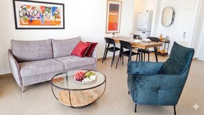 A living room with a grey sofa, a round glass coffee table with fruit, a blue armchair, a dining table with chairs, and a washing machine in the background. - Urban Penthouse Loft W Jacuzzi (Haifa)