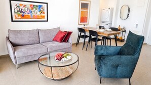 A living room with a grey sofa, a round glass coffee table with fruit, a blue armchair, a dining area with a wooden table, chairs, and a washing machine. - Urban Penthouse Loft W Jacuzzi (Haifa)