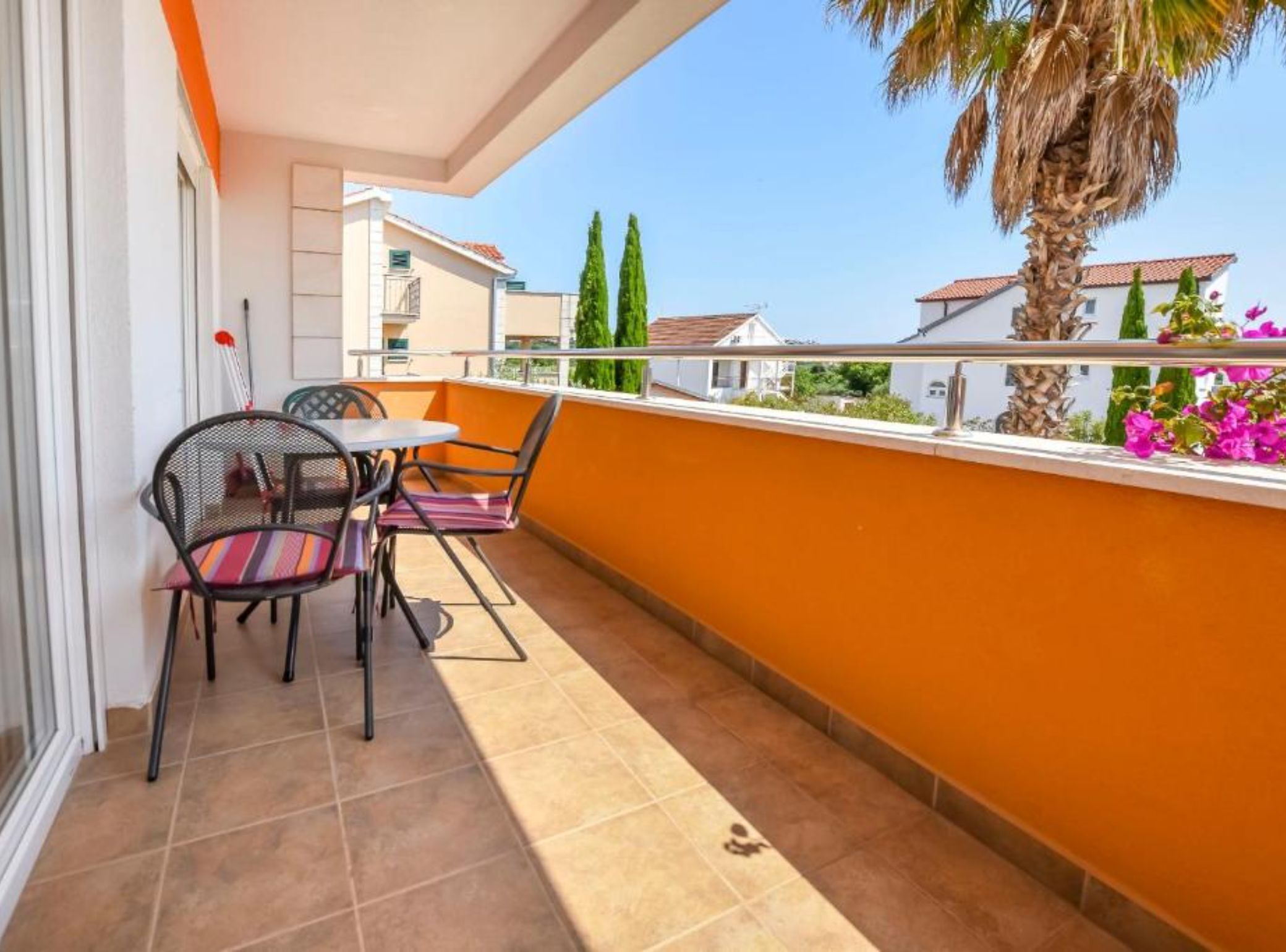 A balcony with a table and chairs, a palm tree, and a view of neighboring buildings.