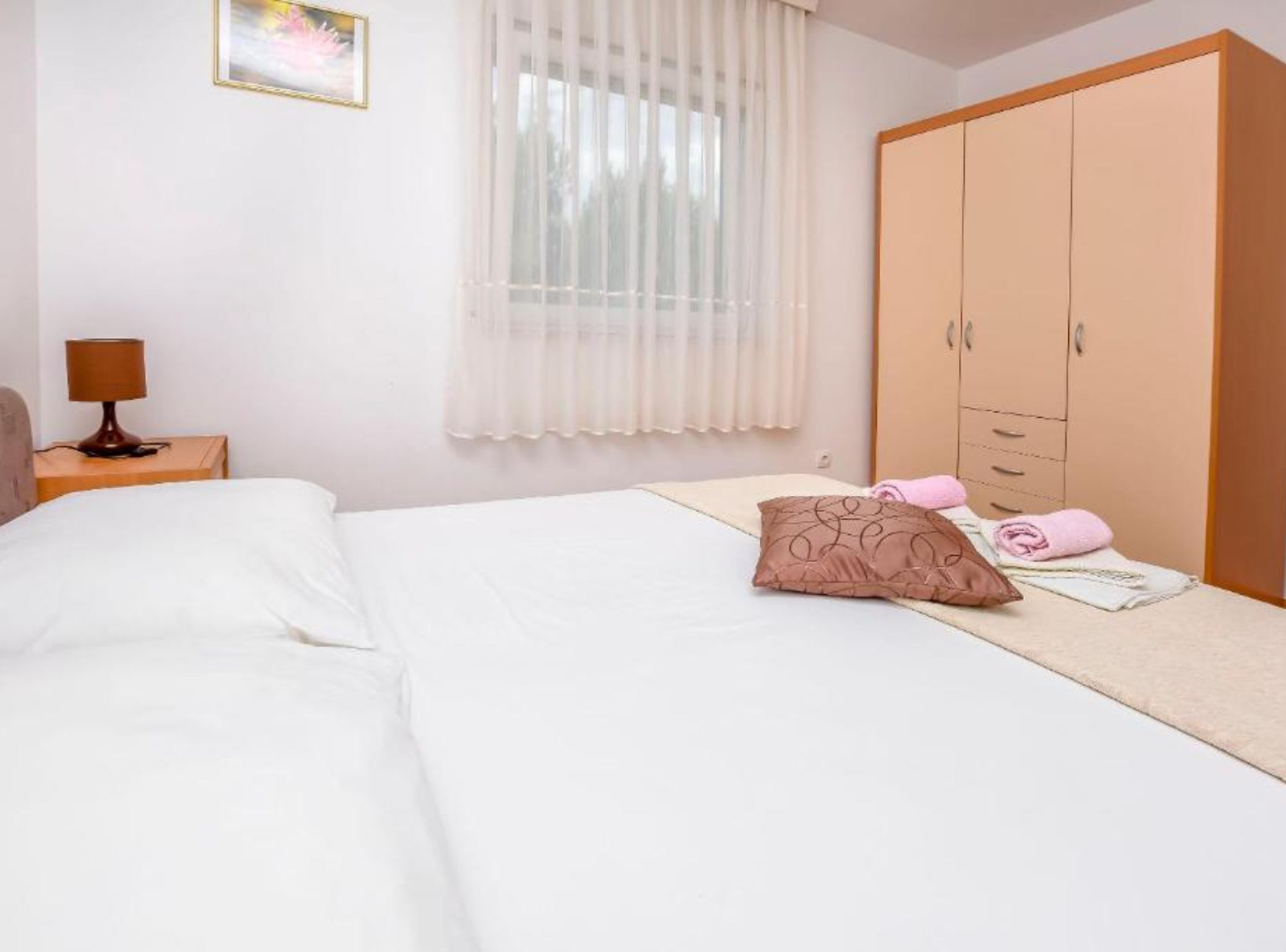 A hotel room with a bed, a nightstand with a lamp, a wardrobe, and a window with sheer curtains.