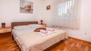 A bedroom with a bed, nightstands, a window with sheer curtains, and a painting on the wall. - Apartments Aurel Suites (Rogoznica)