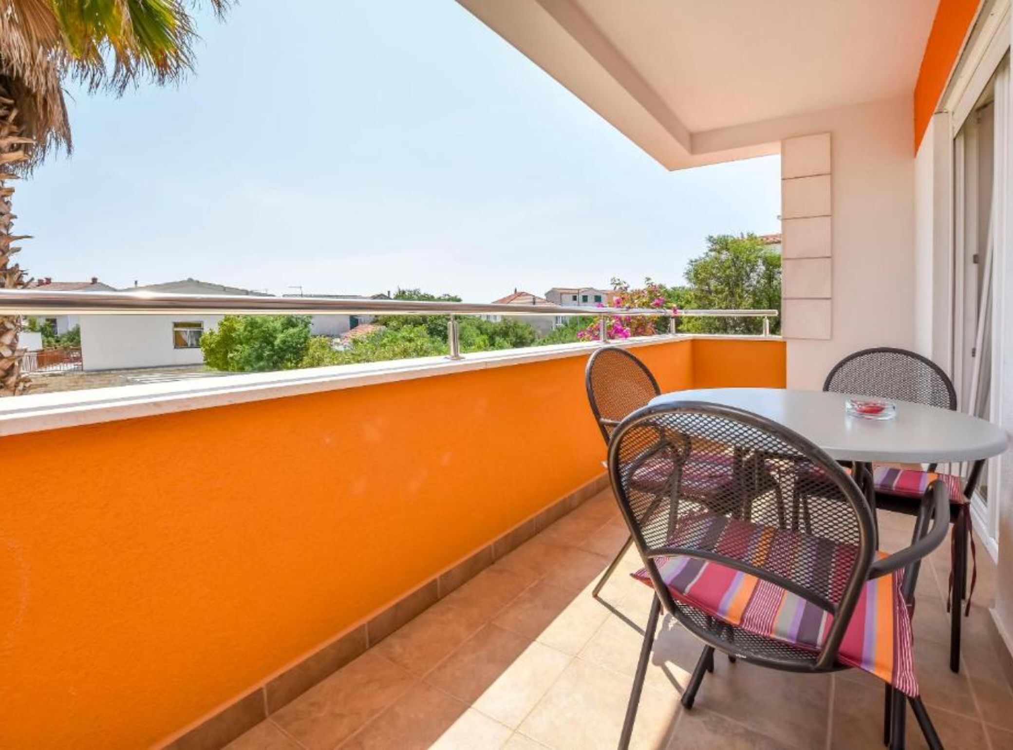 A balcony with orange walls, a table, two chairs, and a view of buildings and greenery.