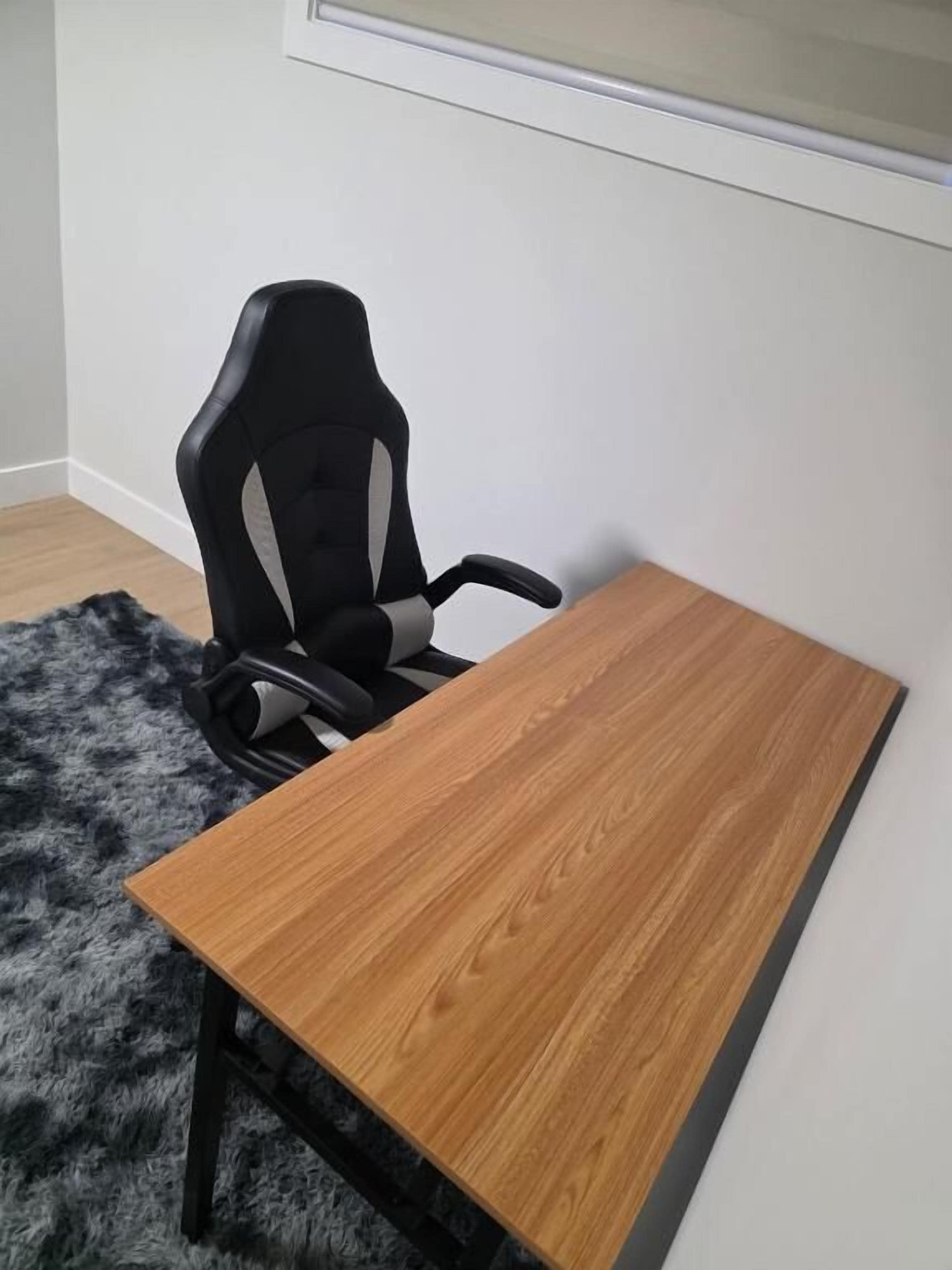 Een kamer met een houten tafel, een zwarte gaming stoel en een pluizig vloerkleed.