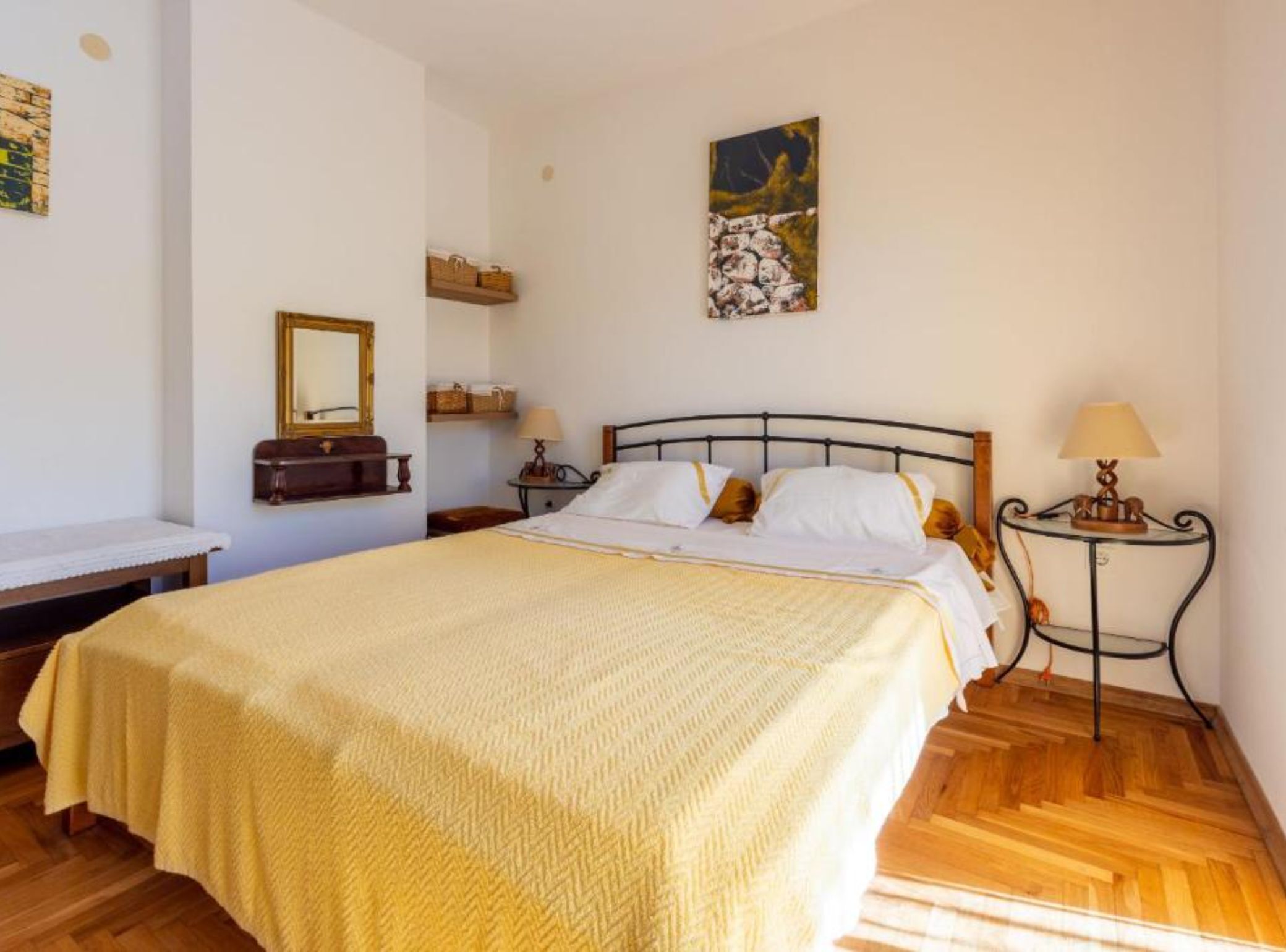 A neatly made bed with a yellow blanket, a bedside table with a lamp, a wall-mounted shelf with baskets, and a painting above the bed.