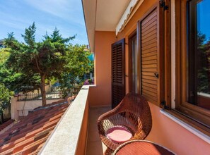 Un balcón con una silla de mimbre, una ventana con contraventanas de madera y un árbol al fondo.