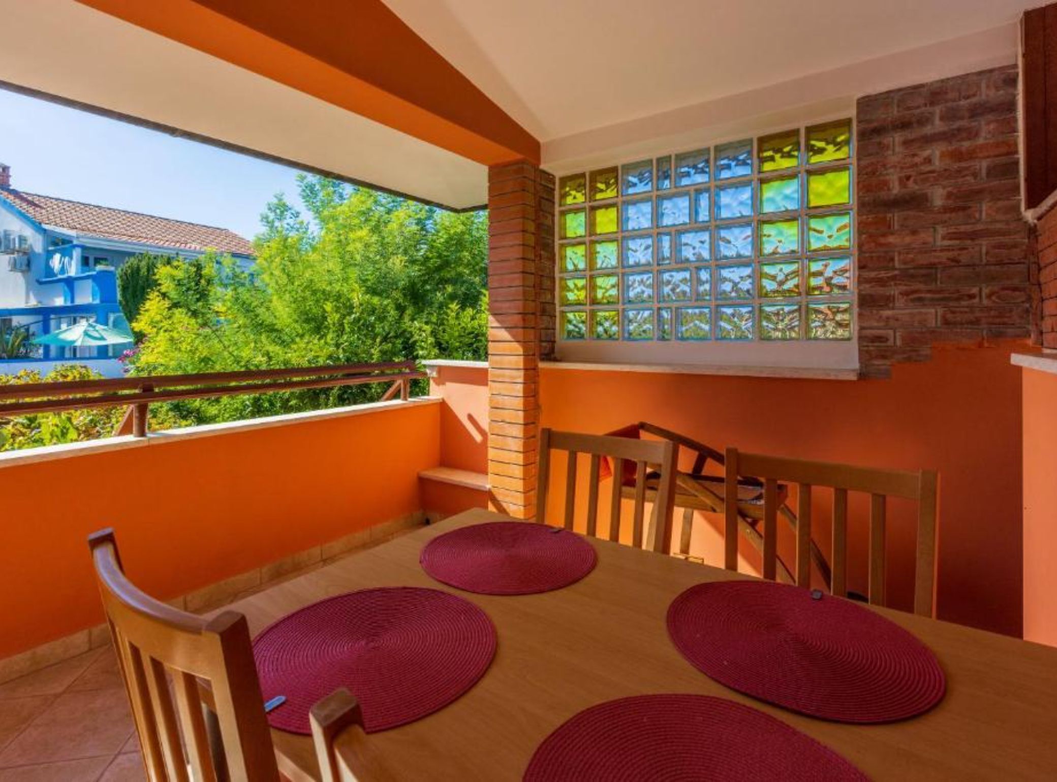 Un balcon avec une table et des chaises en bois, une vue sur la verdure et un immeuble à la façade bleue et blanche.