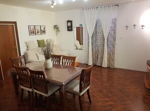 Un comedor con mesa y sillas de madera, un florero con flores, un reloj en la pared y una ventana con cortinas.