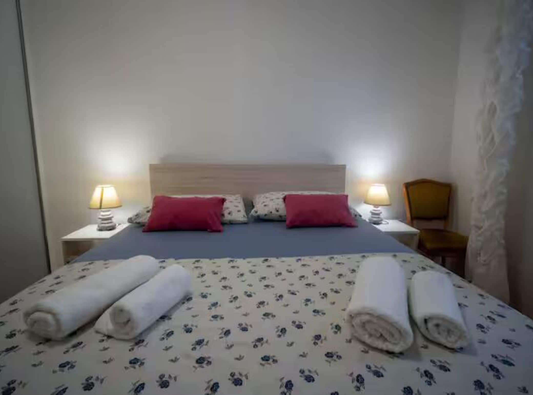 Hotelkamer met een bed, twee kussens, twee bedlampen, een stoel en opgerolde handdoeken op het bed.