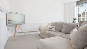 A modern living room with a beige sofa, a painting on the wall, and a window with a view of a building. - New: The Tetbury Suite · 2-bed luxury apt (Tetbury)
