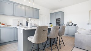 A modern kitchen with grey cabinets, a central island with bar stools, and a built-in oven. - New: The Tetbury Suite · 2-bed luxury apt (Tetbury)