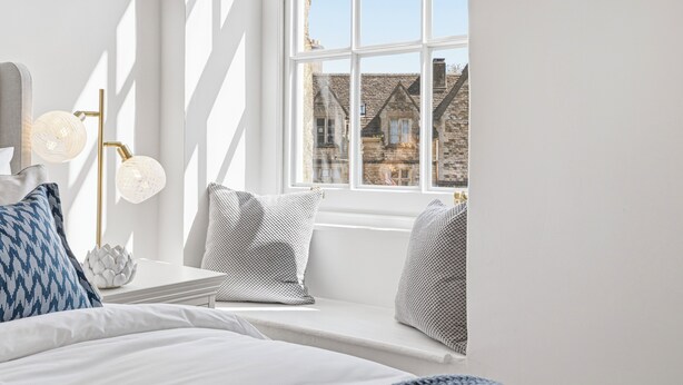 A bedroom with a bed, pillows, a bedside table, a lamp, and a window with a view of buildings. - New: The Tetbury Suite · 2-bed luxury apt (Tetbury)