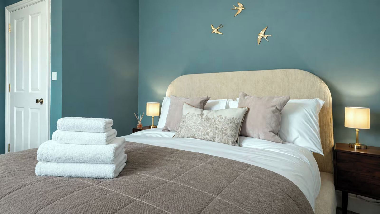 Ein ordentlich bezogenes Bett mit einem beige gefärbten Kopfteil, weißen und beige Kissen sowie einer braunen Decke. Zwei Nachttische mit Lampen, ein Stapel weißer Handtücher und eine Tür mit Messingbeschlägen sind zu sehen.