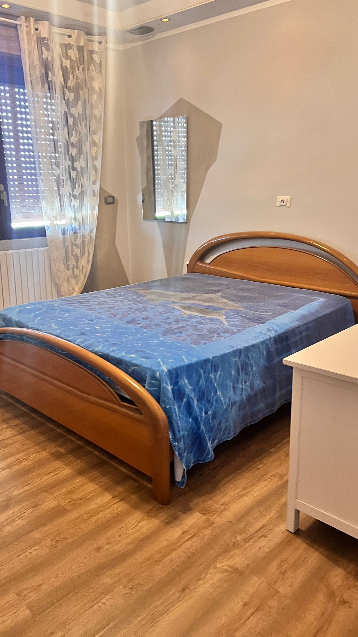 Quarto com estrutura de cama em madeira, capa de colchão azul, mesa de cabeceira e janela com cortinas.