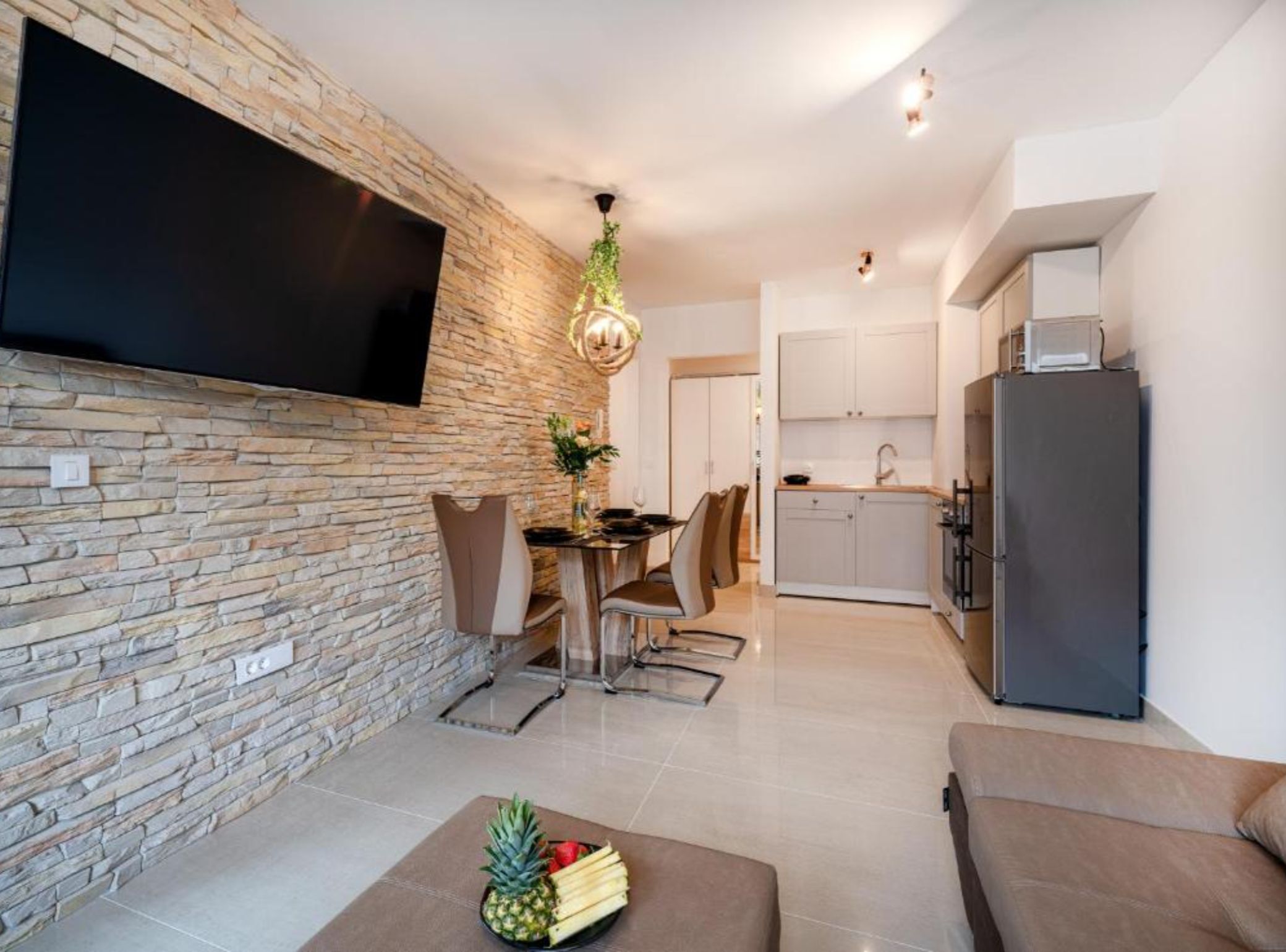 Una sala moderna con pared de piedra, televisor de pantalla plana, zona de comedor con sillas y mesa, cocina con armarios y refrigerador, y un sofá.