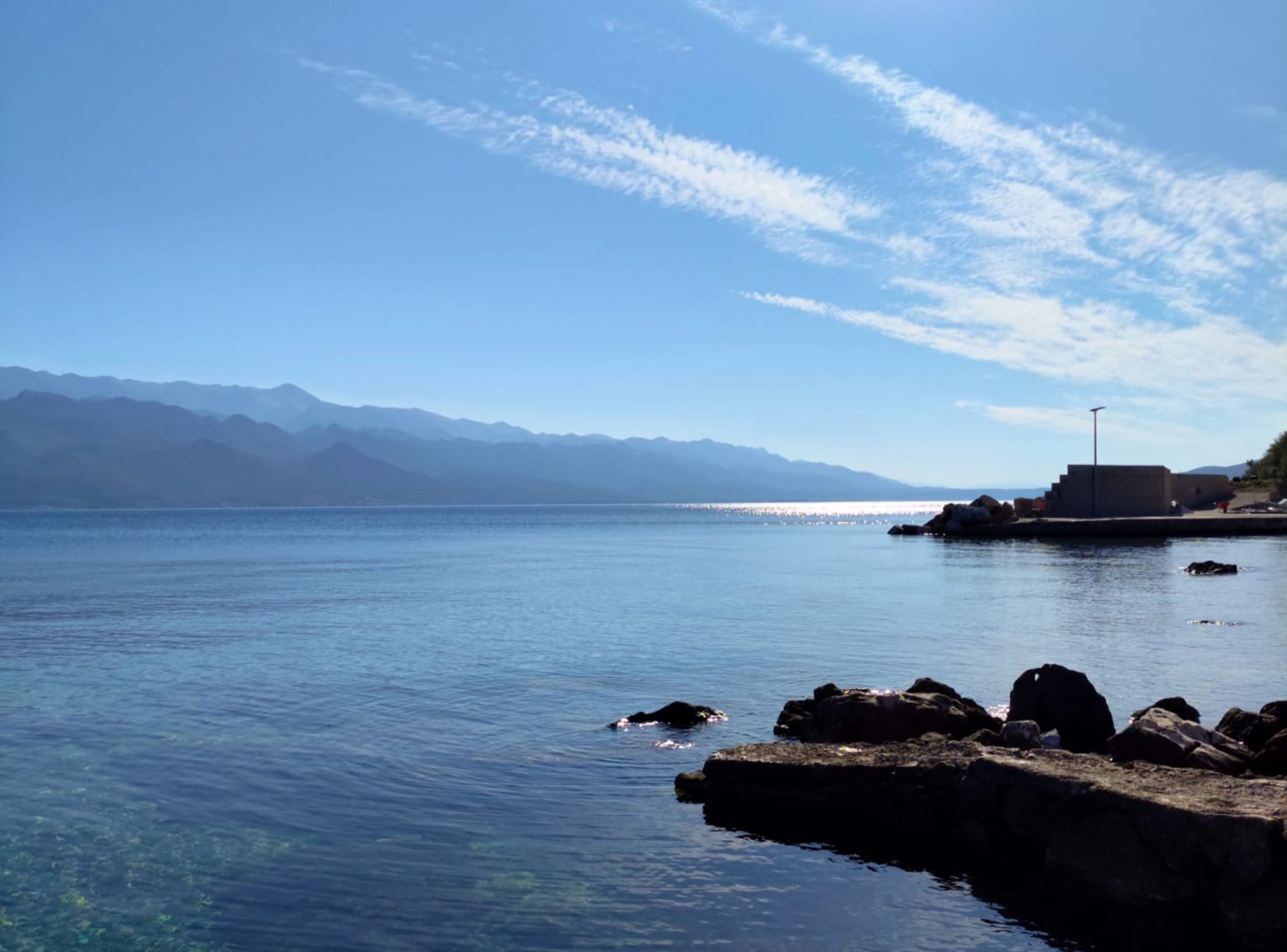 A coastal scene with clear water, a rocky breakwater, and distant mountains under a blue sky.