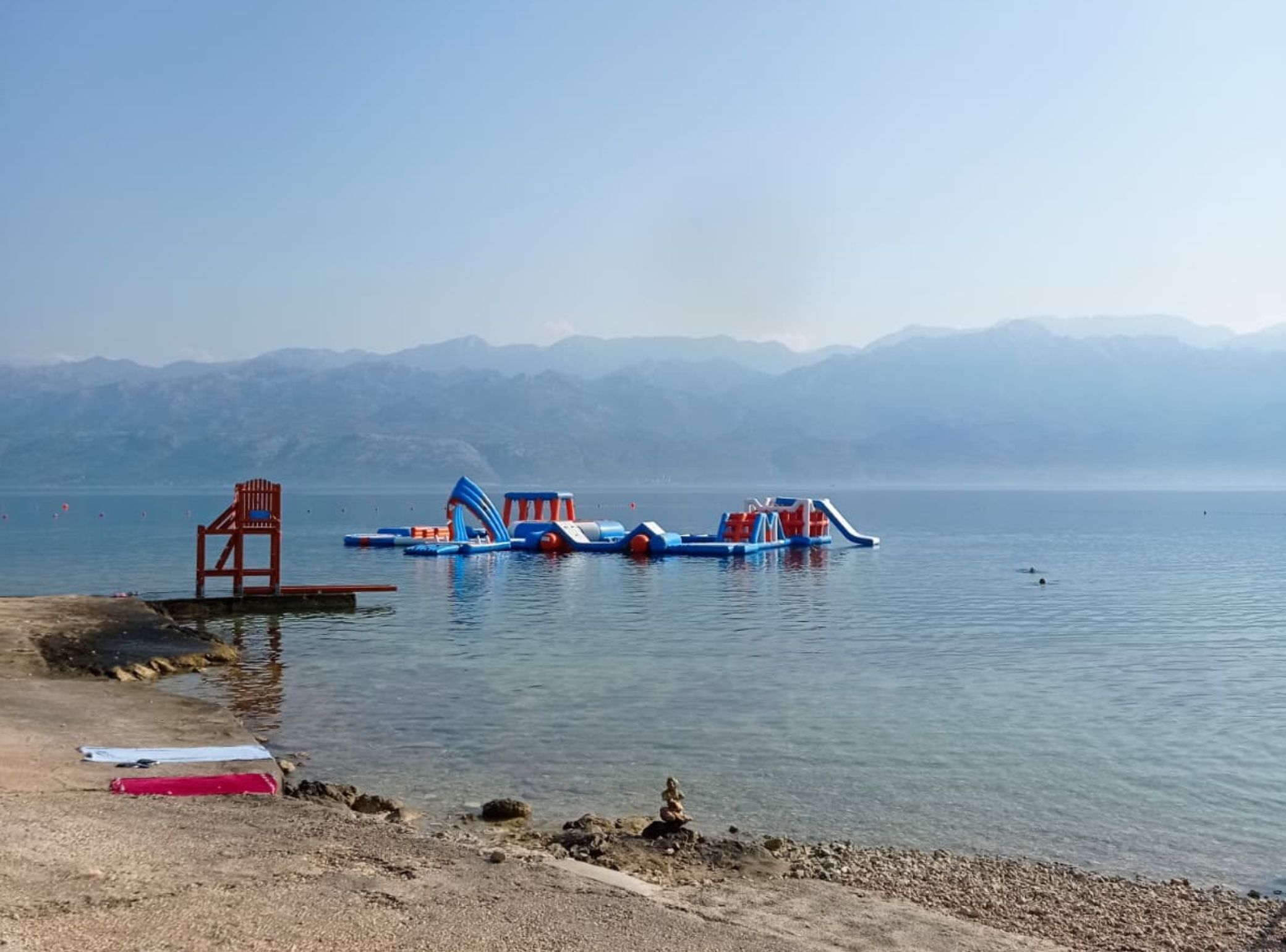 Une plage avec des jeux d’eau gonflables, une chaise rouge et une côte rocheuse.