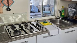 A kitchen with a gas stove, two sinks, a window with curtains, and a view of a living area. - Azzuro Sea Retreat Mobile Home (Razanac)
