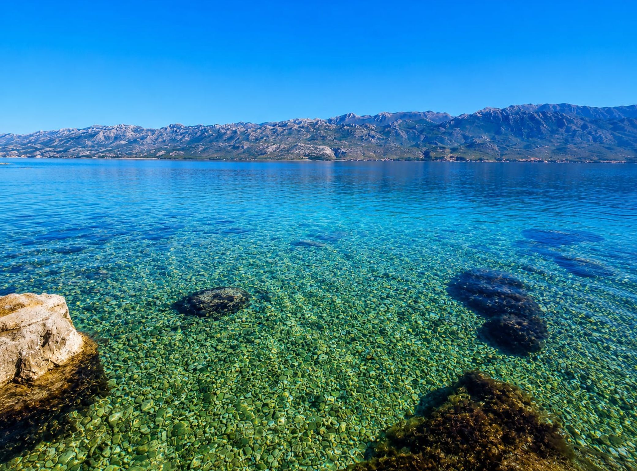 A clear lake with visible pebbles and rocks, surrounded by mountains.