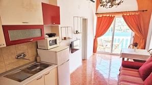 A compact kitchen with a sink, microwave, and refrigerator, a dining area with a table and chairs, and a balcony with orange curtains. - Villa Bonaca (Marina)