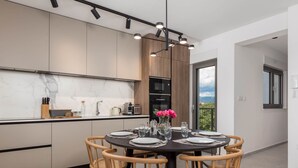 A modern kitchen with a dining area, featuring a round table with chairs, a view to the outside, and built-in appliances. - Apartments Villa Mara With Pool (Omisalj)