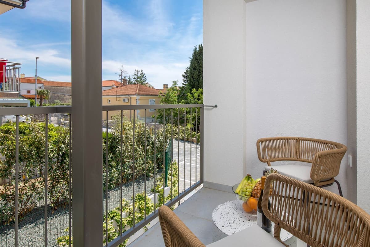 A balcony with wicker chairs, a table with fruit and a bottle, and a view of buildings and greenery.