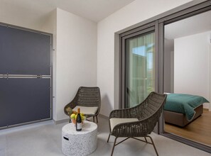 Una habitación moderna con un gran ventanal corredero, dos sillas de mimbre, una mesita redonda con una botella y fruta, y una cama con cubrecama verde.