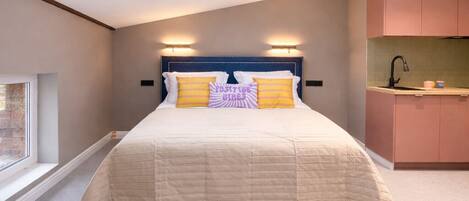 A bedroom with a skylight, a bed with a beige quilt, and a kitchenette with pink cabinets.