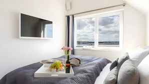 A bedroom with a bed, a bedside table with a tray of fruit and flowers, a window with a view of a cityscape, and a mounted TV. - Nothing Beats the Sea – 2-Bedroom Apartment for 5 with Harbor View in Kappeln (Kappeln)