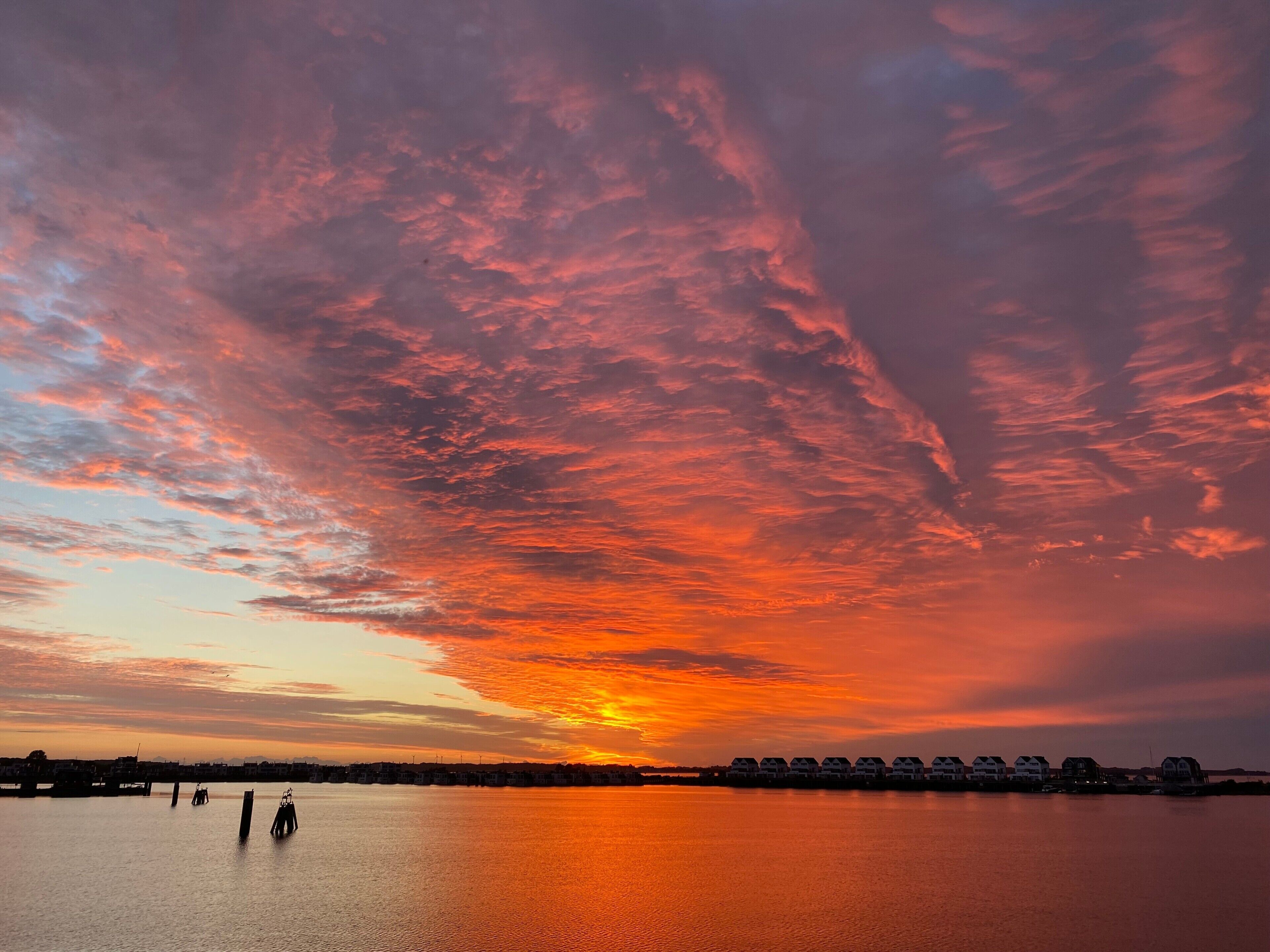 Un coucher de soleil sur une étendue d’eau calme, avec une rangée de maisons le long de la rive.