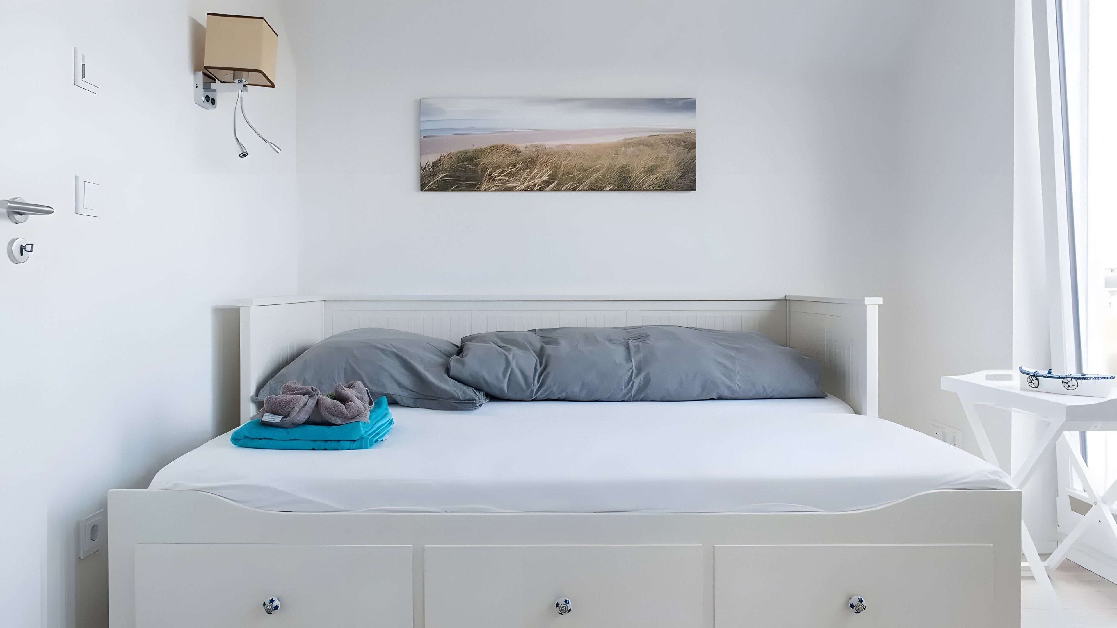Ein Bett mit weißem Kopfteil, grauer Bettwäsche und einem Landschaftsbild darüber.