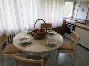Et spiseområde med et rundt bord, fire stole, en kurv med frugt og en komfur i baggrunden.