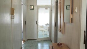 Um corredor com porta que dá para uma varanda, uma pequena mesa com um cesto, e um relógio na parede.