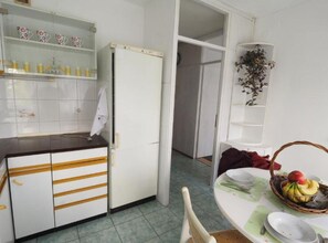 Et køkken med et køleskab, et spisebord med en kurv med frugt og en plante på en hylde.